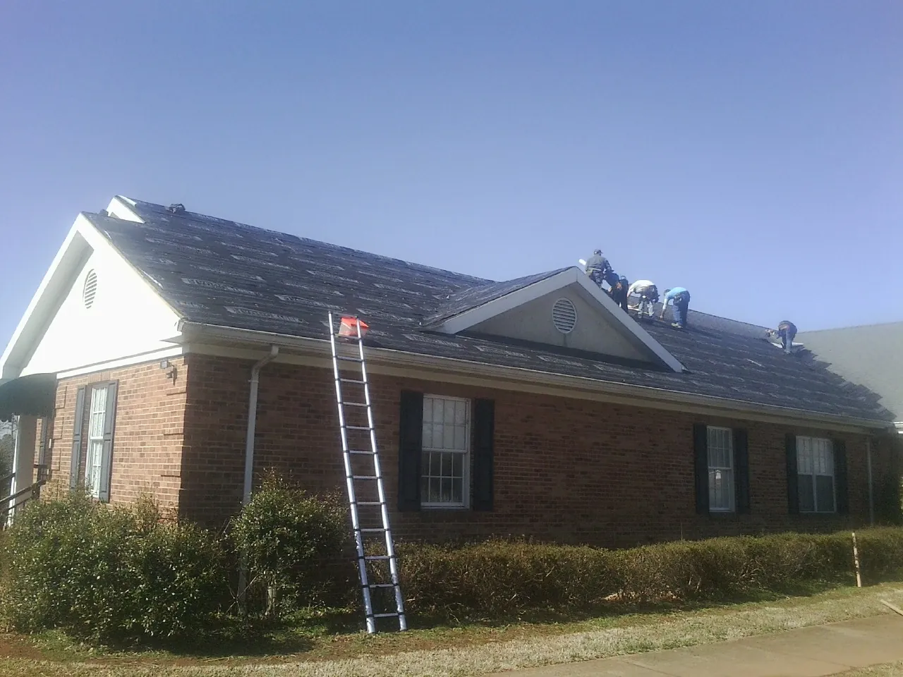 Crew tearing shingles off a brick ranch roof with a ladder set — Emergency Roof Repair work by our crew in Fort Covington