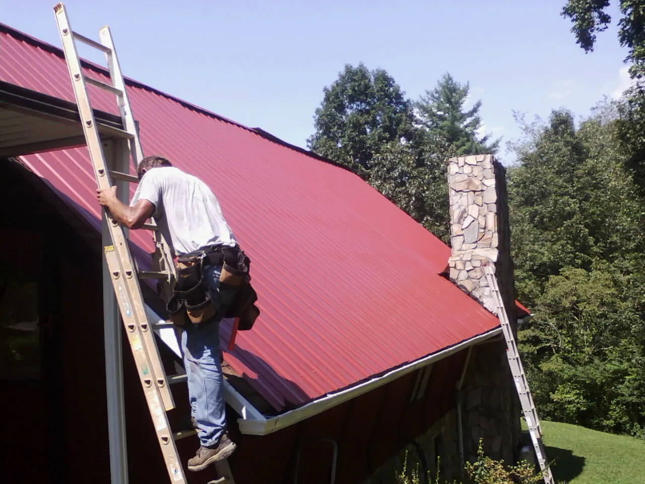 Roofer on a ladder beside a red metal roof and stone chimney — Soffit & Fascia Repair by Roofing Brothers in Fort Covington